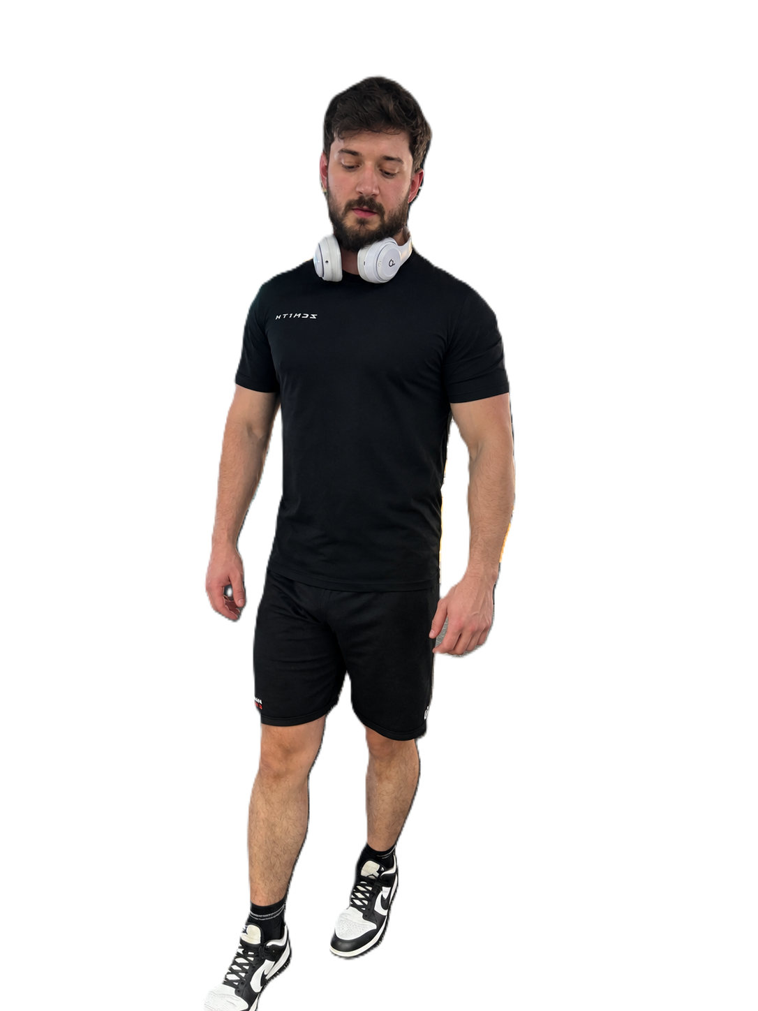 A man wearing a black athletic t-shirt and matching black shorts, with white headphones around his neck and black-and-white sneakers, standing against a plain white background.