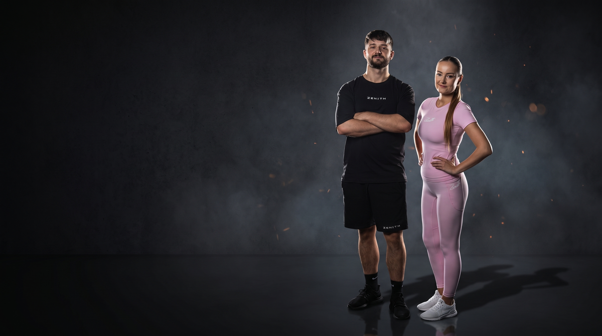 A man and a woman standing in a dark studio setting, wearing athletic apparel. The man wears a black short-sleeve top and black shorts, while the woman wears a matching light pink short-sleeve top and leggings.