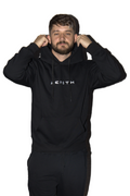 A man wearing a black pullover hoodie with 'ZENITH' printed in white across the chest, adjusting the hood. He is also wearing black sweatpants against a solid white background.