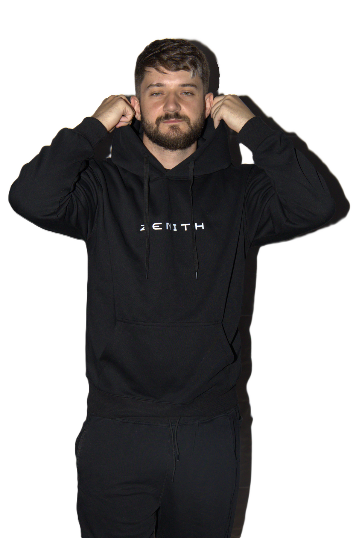 A man wearing a black pullover hoodie with 'ZENITH' printed in white across the chest, adjusting the hood. He is also wearing black sweatpants against a solid white background.