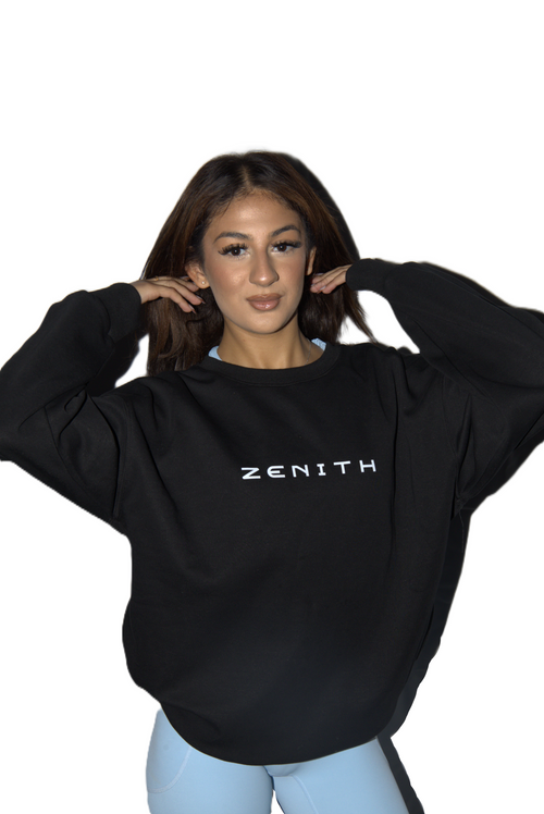 A woman wearing a black crewneck sweatshirt with the brand name 'ZENITH' printed in white across the chest, paired with light blue leggings, set against a solid white background.
